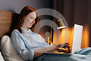 Side view of redhead young woman using laptop computer lying on bed at cozy dark bedroom.