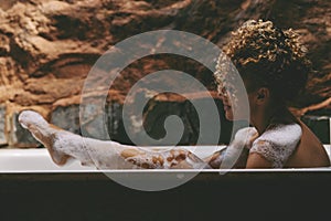 Side view of relaxed african american naked young woman taking bath at home, laying in beautiful bathtub, rubbing her skin with