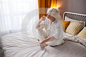 Side View On Relaxed Happy Elderly Female Sitting On Bed And Drinking Water, Looking At Side At Window