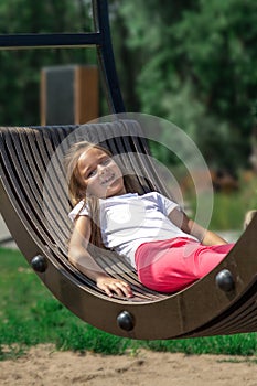 Side view of relaxed pensive little girl in casual clothes lying on a swing and spending a sunny summer day in the park