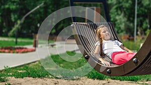 Side view of relaxed pensive little girl in casual clothes lying on a swing and spending a sunny summer day in the park. Girl is