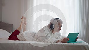 Side view relaxed slim mature woman lying on bed in headphones listening to music on tablet playlist. Happy carefree