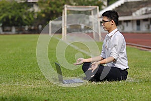 Side view of relaxed young Asian business man with laptop doing yoga position on the green grass of stadium.