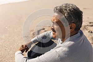 Side view of retired biracial senior man looking away while sitting at beach on sunny day