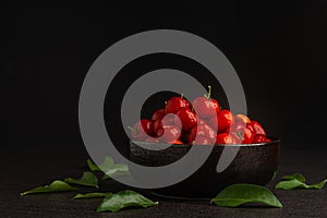Side view of ripe red acerola cherries fruit in a ceramic bowl with a black background.