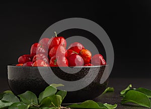 Side view of ripe red acerola cherries fruit in a ceramic bowl with a black background.