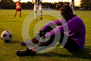 Side view of sad african american male goalkeeper sitting on field at playground during sunset