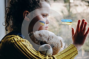 Side view of sad little girl kid with flag of ukraine on face, tired child holding fluffy bear sitting alone in windowsill at home