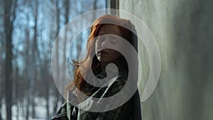 Side view of sad young woman sitting by window on sunny day on windowsill with winter nature background, feels upset