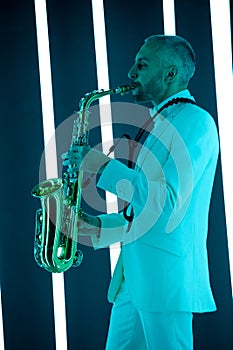 Side view saxophone player in a white suit against the backdrop of neon lamps with blue backlight. Saxophonist jazzman