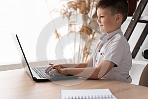 Side view of a schoolboy at home schooled working on a laptop typing text on the keyboard while communicating with a photo