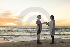 Side view of senior biracial bride putting wedding ring on groom\'s finger at beach during sunset