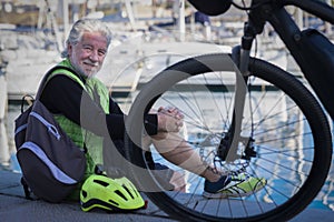 Side view of a senior man with white hair sitted at port resting near his bicycle. Sailboats in the background, blue sea. Happy