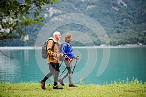 A side view of senior pensioner couple hiking by lake in nature.