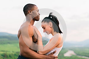 Side view of fit mixed race couple with perfect bodies in sportswear softly embracing on mountains landscape