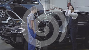 Side view of shocked Caucasian man standing next to his broken car as auto mechanic coming up. Desperate car owner