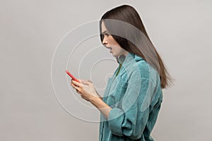 Side view of shocked pretty woman reading message on smartphone with open mouth, using mobile device