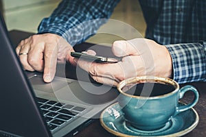 Side view shot of busy businessman man`s hands using smart phone in interior and laptop at office desk.