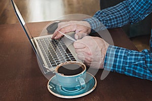 Side view shot of a man`s hands holding card and using laptop sitting at wooden table with cup of coffee. Close up.