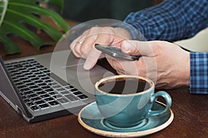 Side view shot of a man`s hands using smart phone and laptop sitting at wooden table. Close up.