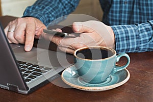 Side view shot of a man`s hands using smart phone and laptop sitting at wooden table with cup of black coffee. Close up.