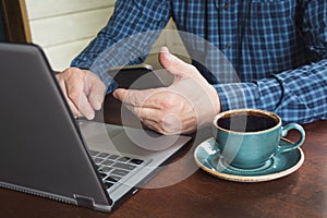 Side view shot of a man`s hands using smart phone and laptop sitting at wooden table with cup of black coffee. Close up.