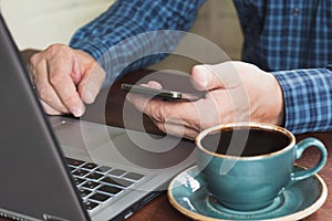 Side view shot of a man`s hands using smart phone and laptop sitting at wooden table with cup of coffee. Close up.