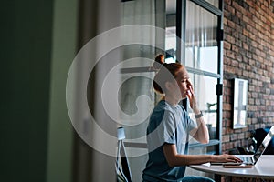 Side view of sleepy redhead young woman yawning while working on laptop computer sitting at table