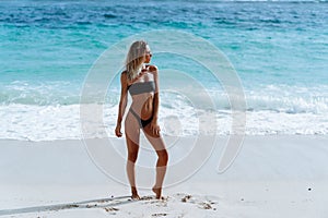 Side view of slim sexy model posing in swimsuit and sunglasses background of ocean on beach at sunny day