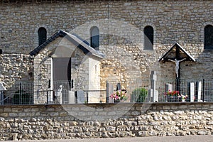 Side view of a small church with wooden door, stone walls, windows, tombs, a cross with Jesus surrounded by a metal fence