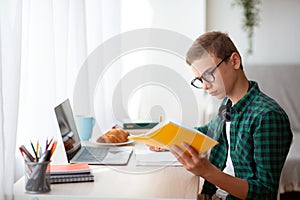 Side view of smart schooler reading book and using laptop