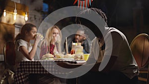 Side view of smiling African American young man texting on smartphone as blurred family talking laughing eating picnic