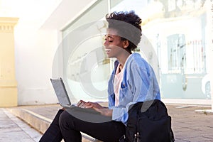 Side view smiling african female student sitting on sidewalk with laptop