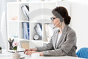 Side view of smiling attractive call center operator in glasses and headset using computer at table