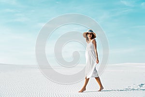 View of smiling beautiful girl in white dress and straw hat walking on sandy beach with blue sky