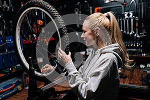 Side view of smiling blonde female cycling craftsman checking bicycle wheel spoke with bike spoke key in repair workshop