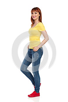 Side View Of Smiling Casual Young Woman Standing With Hands In Pocket And Looking At Camera