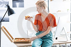 side view of smiling female freelancer sitting on table and eating pizza at home photo