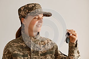 Side view of smiling female soldier wearing military costume, posing indoor in light room, standing with key in hands, buying new