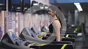 Side view of smiling fit Middle Eastern young woman walking on treadmill in gym. Portrait of confident slim beautiful