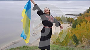 Side view smiling happy woman in national Ukrainian embroidered dress stretching flag standing at background of autumn