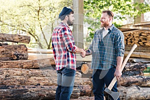 side view of smiling lumberjack with tattooed hand holding axe and shaking hands with partner