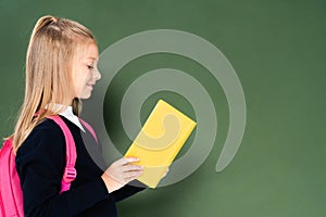 side view of smiling schoolgirl reading book while standing near green chalkboard.