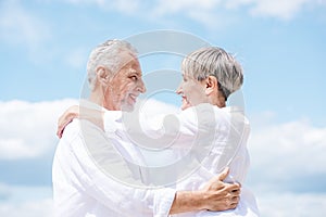 side view of smiling senior couple embracing and looking at each other under blue sky.