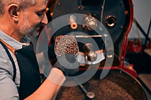 Side view smiling unshaved master smelling coffee grains while keeping them in ladle. He standing near industrial technical