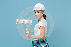 Side view of smiling woman in protective helmet hardhat hold paint roller for wall painting isolated on blue background