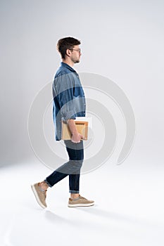 Side view of a smiling young casual man walking, student with book ang notes on white background.