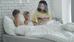 Side view of sons lying in bed and listening to mother reading book. Portrait of happy African American family resting