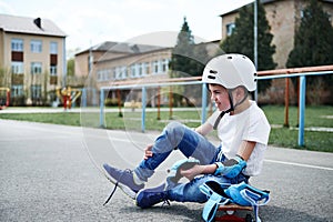 Side view of a sporty boy in safety helmet sitting on skateboard and putting on protective knee pads