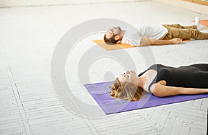 Side view sporty woman and man practicing yoga together, lying in resting pose Savasana in white yoga studio.